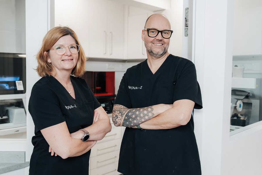 Eine Zahntechnikerin und ein Zahntechniker im praxiseigenen Dentallabor von NOVA–Z. Der Hintergrund ist hell und freundlich mit hochmoderner Technik. Die beiden lächeln sympathisch in die Kamera