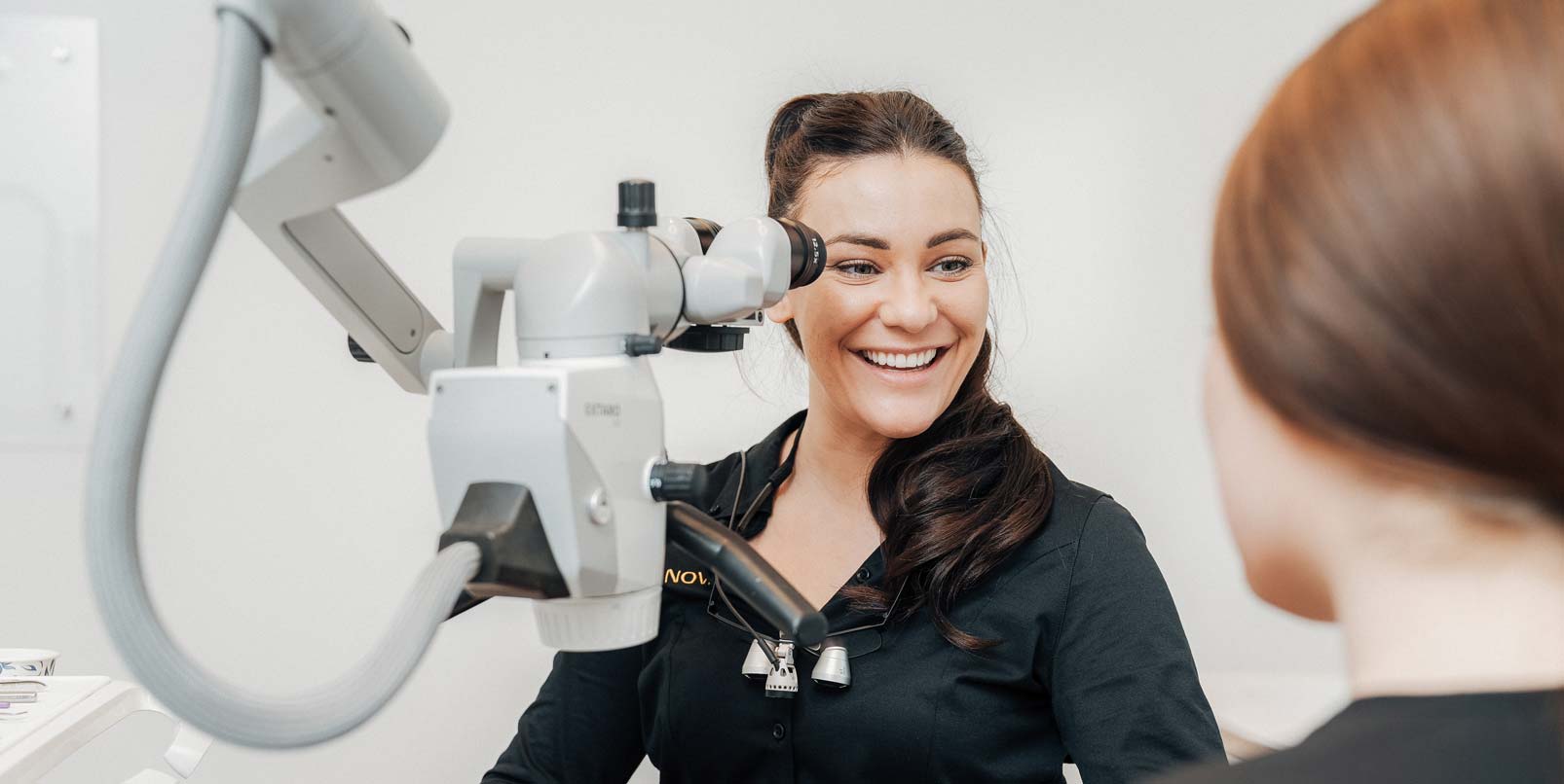 Zahnärztin Theresa Spieß von NOVA-Z in München Harlaching sitzt vor einem Dentalmikroskop und lächelt sehr sympathisch ihre Kollegin an