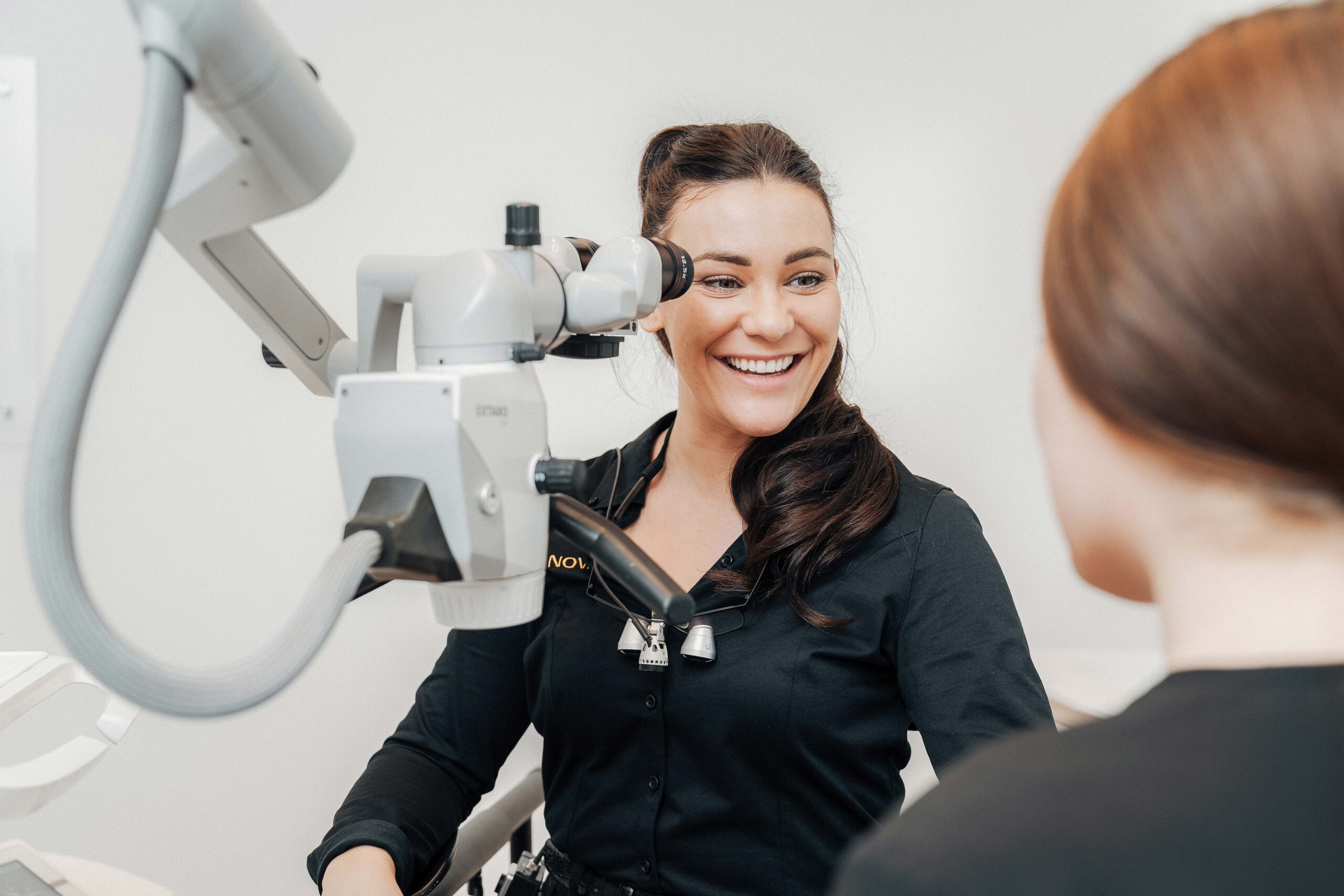 Zahnärztin Theresa Spieß lächelt sehr freundlich und sympathisch ihre Kollegin an. Sie sitzt vor einem Dentalmikroskop.