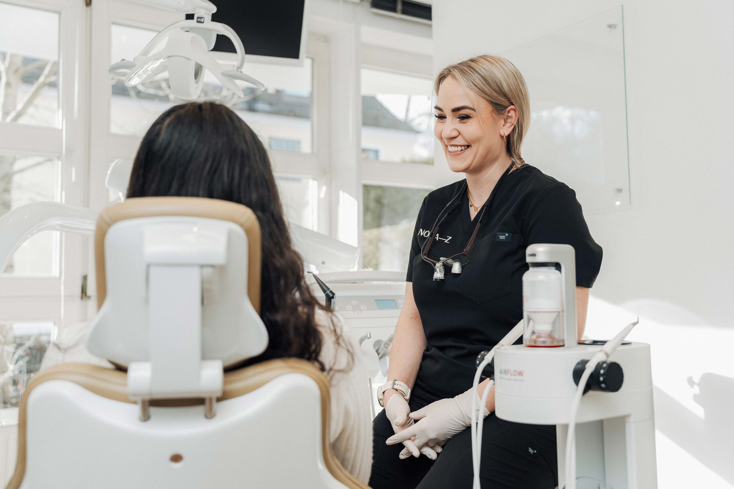 Eine Dentalhygienikerin von NOVA–Z spricht sehr sympathisch und freundlich lächelnd mit einer Patientin. Die Praxis ist lichtdurchflutet und freundlich mit großen Fenstern im Hintergrund.