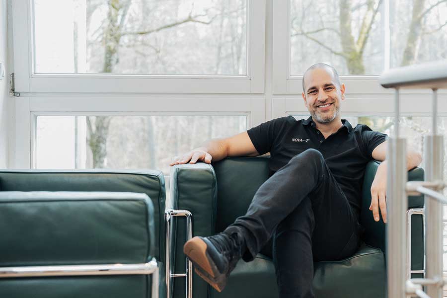 Zahnarzt Dr. Denis Novakovic sitzt in einem dunkelgrünen Ledersessel vor einer hellen Fensterfront in seinem Zentrum für innovative Zahnmedizin NOVA–Z in München Harlaching. Er lacht sympathisch in die Kamera.