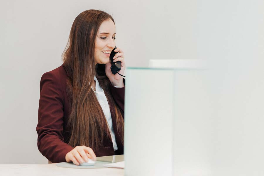 Eine Mitarbeiterin am Empfang telefoniert sympathisch lächelnd mit einer Patientin und schaut dabei auf einen Bildschirm, mit der Hand an der Computermaus.