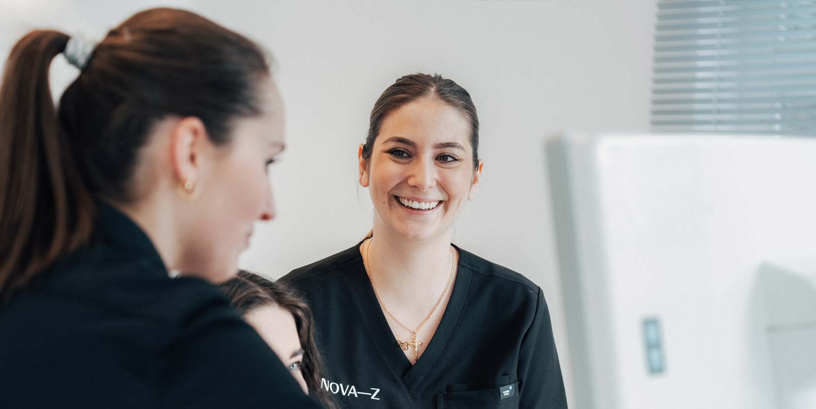 Eine zahnmedizinische Fachangestellte lächelt während der Beratung einer Patientin fröhlich die Zahnärztin an. Beide tragen schwarze Praxiskleidung mit dem Logo NOVA-Z