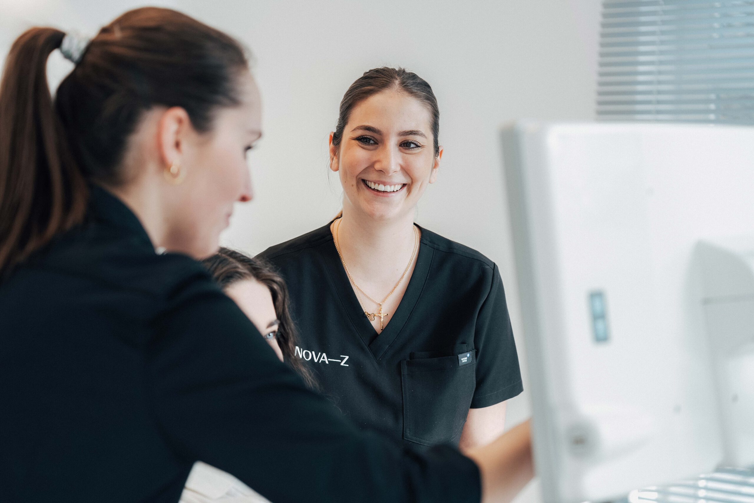 Eine zahnmedizinische Fachangestellte lächelt während einer Patientenberatung die Zahnärztin strahlend an, während die Zahnärztin gerade etwas mit der Patientin am Bildschirm bespricht.