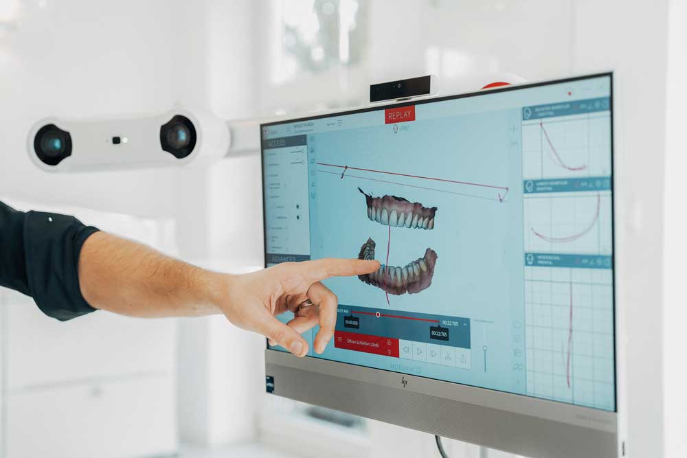 Dr. Denis Novakovic zeigt mit seinem Finger auf einen Bildschirm, auf dem eine 3D-Grafik eines Gebisses und Kieferbewegungsanalysen und Kiefergelenksdaten in Echtzeit zu sehen sind.