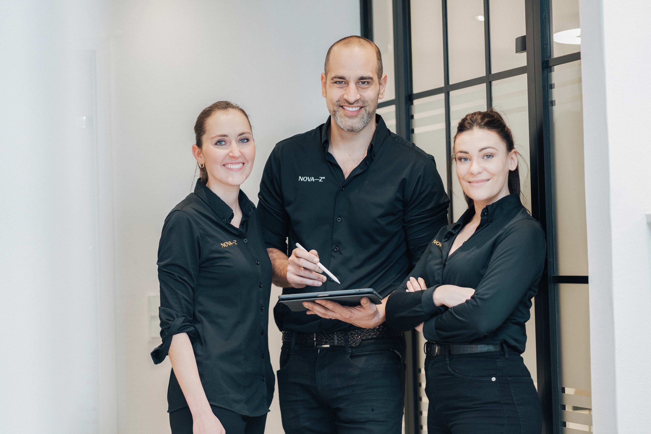 Dr. Denis Novakovic zusammen mit Dr. (Uni Budapest) Nathalie Schubert und Zahnärztin Theresa Spiess. Alle drei tragen schwarze Kleidung mit dem Logo NOVA–Z und lächeln sehr freundlich und sympathisch in die Kamera.