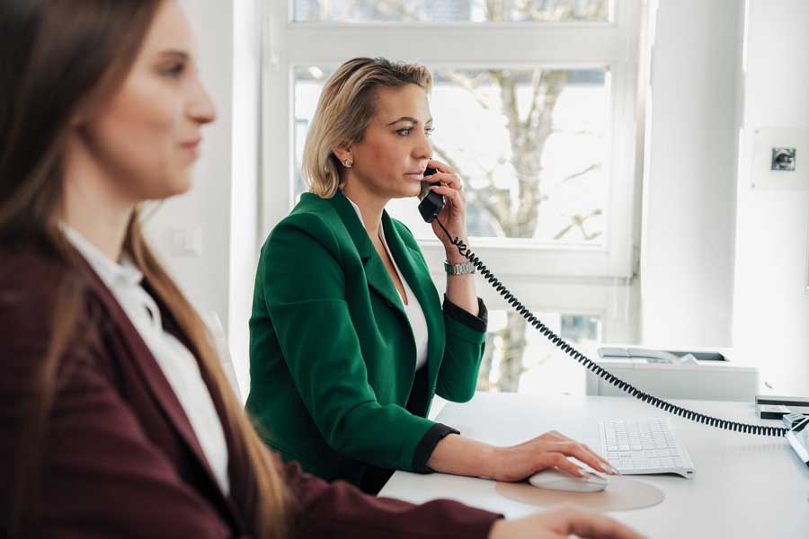 Eine Mitarbeiterin von NOVA–Z am Empfang telefoniert und gibt an einem Bildschirm etwas ein, eine zweite Mitarbeiterin ist unscharf im Vordergrund zu sehen.