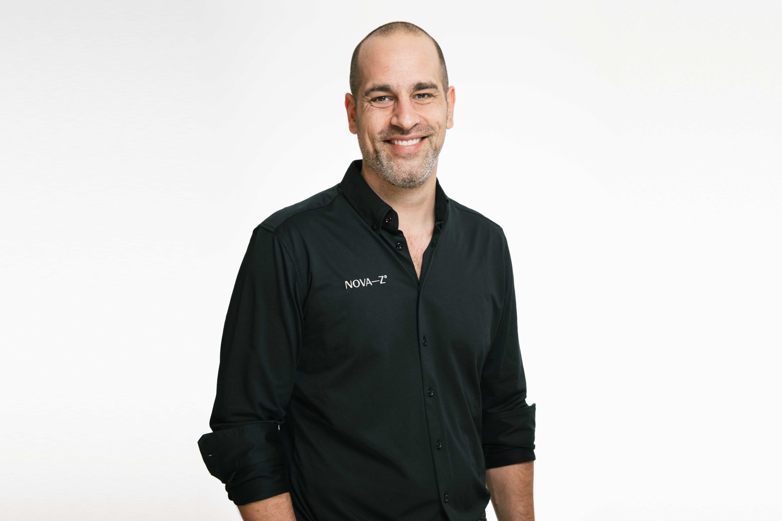 Porträtbild von Zahnarzt Dr. Denis Novakovic vor weißem Hintergrund. Er trägt ein schwarzes Hemd bestickt mit dem weißen Logo NOVA-Z und lächelt freundlich und sehr sympathisch in die Kamera