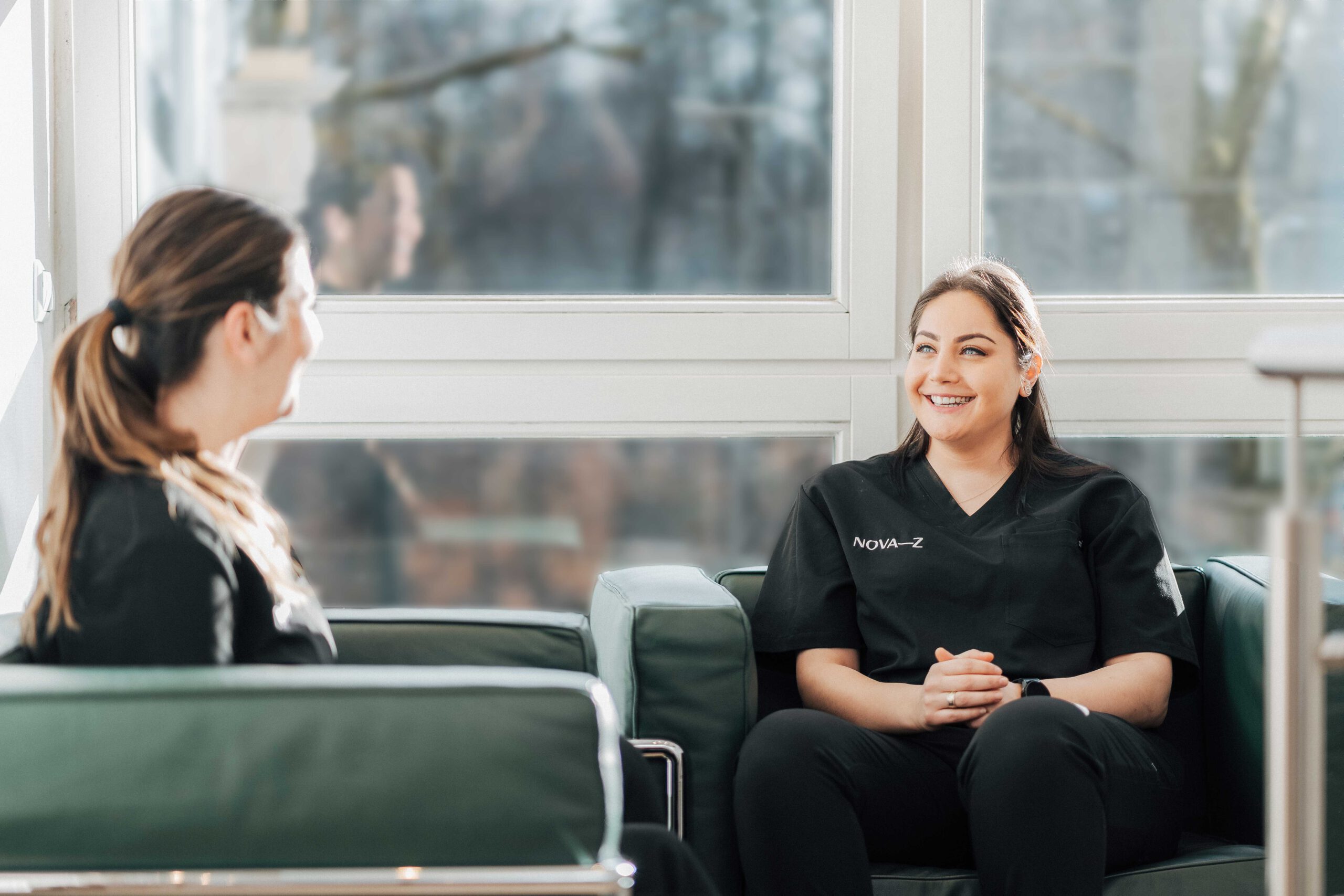 Zwei zahnmedizinische Fachangestellte sitzen in dunkelgrünen Ledersesseln in der modernen Zahnarztpraxis und lachen sich sympathisch und freundlich an. Beide tragen dunkle Praxiskleidung bestickt mit dem Logo NOVA–Z in weiß.