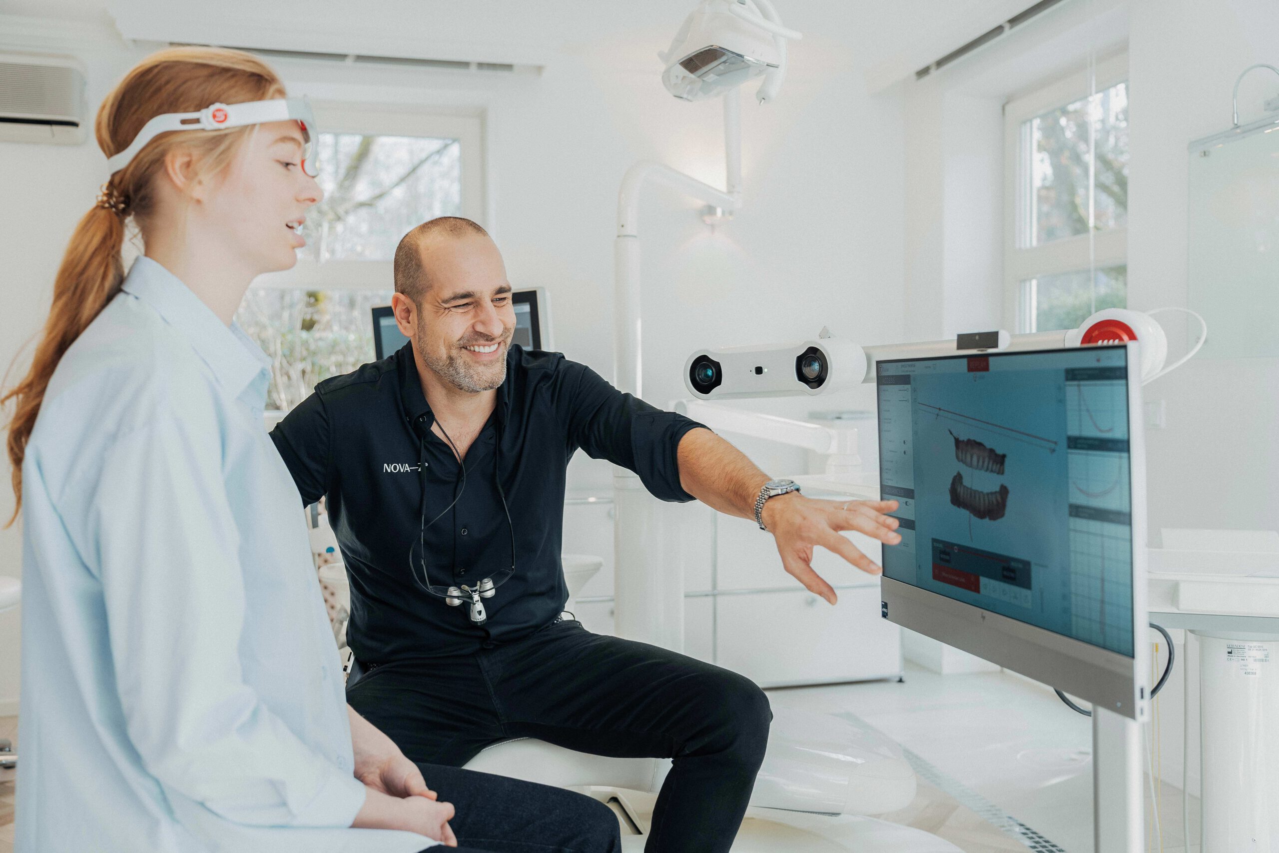 Zahnarzt Dr. Denis Novakovic bespricht sympathisch lächelnd mit einer Patientin an einem Bildschirm 3D-Bilder ihres Mundraums