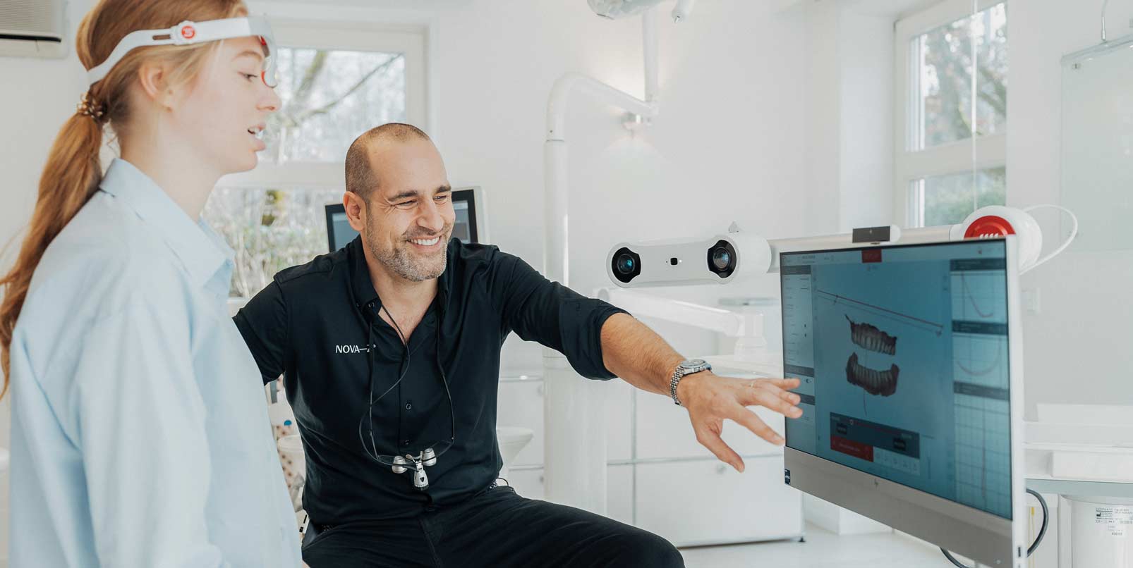Zahnarzt Dr. Denis Novakovic bespricht mit einer Patientin an einem Bildschirm ihr Gebiss. Er lächelt sehr freundlich dabei.