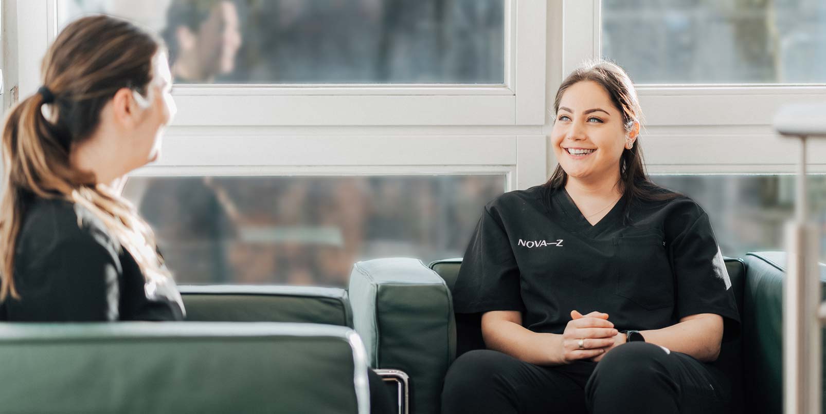 Zwei zahnmedizinische Fachangestellte von NOVA-Z sitzen in grünen Ledersesseln und lachen vergnügt. Beide tragen schwarze Praxiskleidung mit dem Logo NOVA–Z.