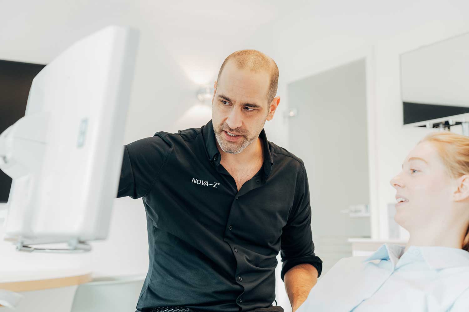Zahnarzt Dr. Denis Novakovic erklärt einer Patientin im Behandlungsstuhl etwas an einem Bildschirm. Dabei zeigt er mit dem Finger auf das Display